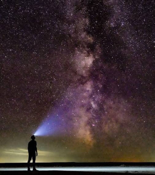 Una persona in silhouette sulla riva illumina la Via Lattea e il cielo stellato con una lampada frontale brillante.