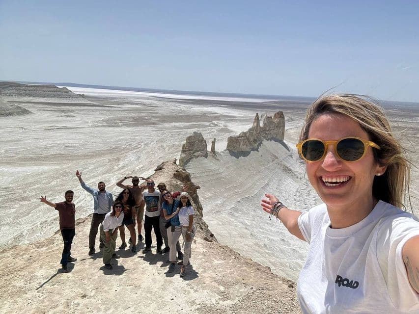 Una donna si scatta un selfie sorridente con un gruppo WeRoad, in posa su una cresta rocciosa che domina un vasto paesaggio bianco e arido sotto un cielo sereno.