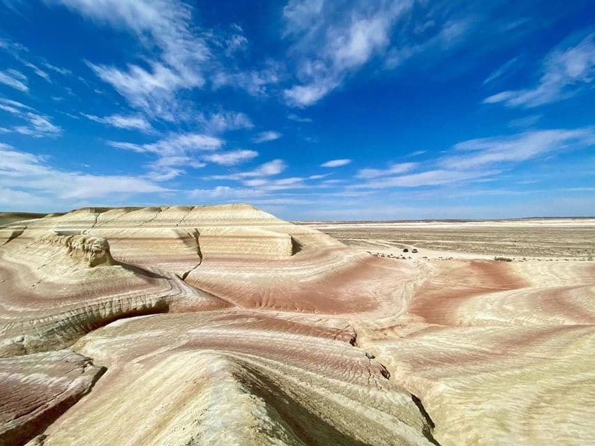 Formazioni rocciose colorate e stratificate si estendono in un deserto sotto un cielo azzurro brillante con nuvole e due veicoli in lontananza.