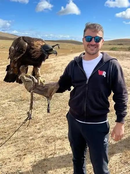 Un uomo sorridente con una felpa WeRoad nera e occhiali da sole tiene un'aquila reale incappucciata sul braccio guantato in un campo erboso.
