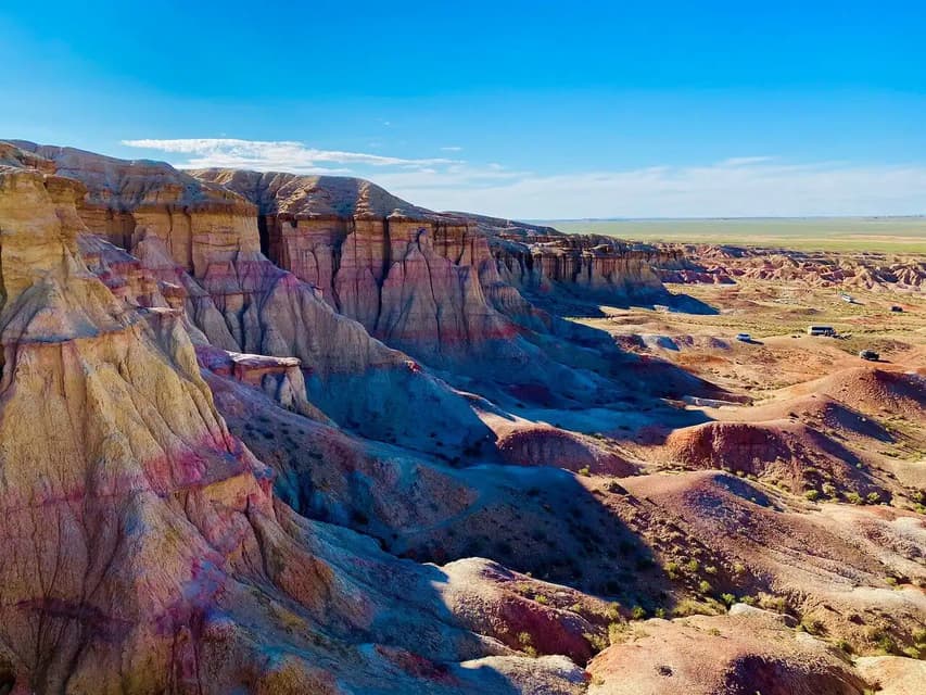 Un vasto canyon desertico con formazioni rocciose colorate e stratificate sotto un cielo azzurro brillante, con veicoli turistici visibili in lontananza.