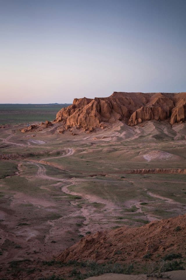 Un vasto paesaggio desertico con grandi formazioni rocciose rosse e tortuosi sentieri sterrati sotto un cielo pallido al tramonto.