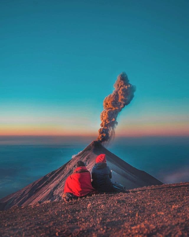 Two people sit on a mountainside watching an erupting volcano send a plume of smoke into the sunrise sky.