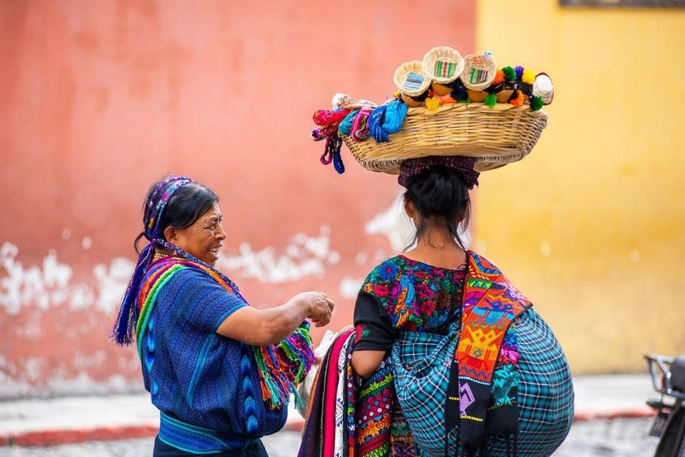 Two women in colorful traditional clothing on a street, one balances a woven basket full of goods on her head.