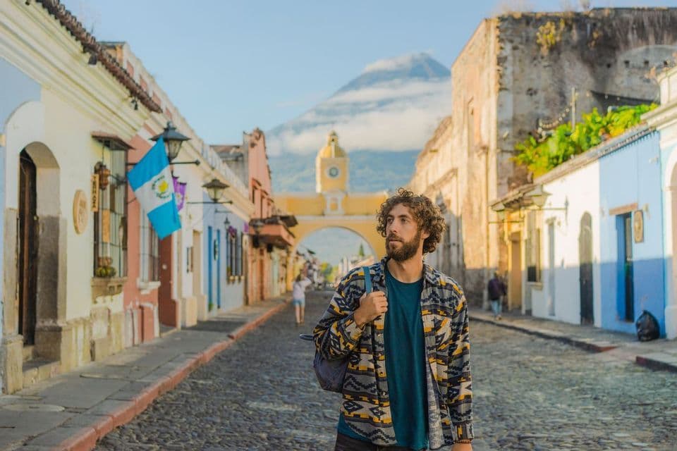 A man with a backpack stands on a cobblestone street lined with colorful buildings, with a yellow arch and a large volcano in the background.