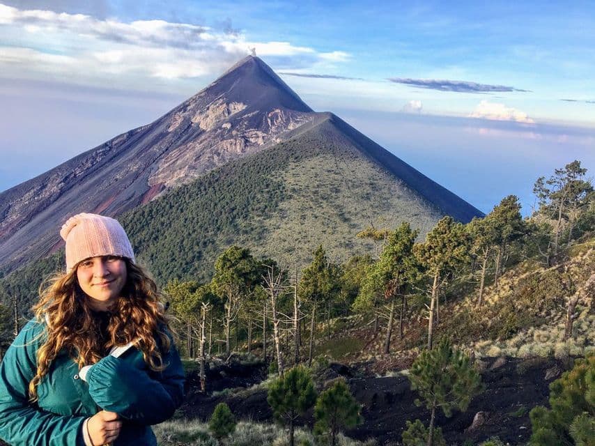 A woman in a pink beanie and green jacket smiles on a mountainside, with an active volcano erupting smoke in the background.