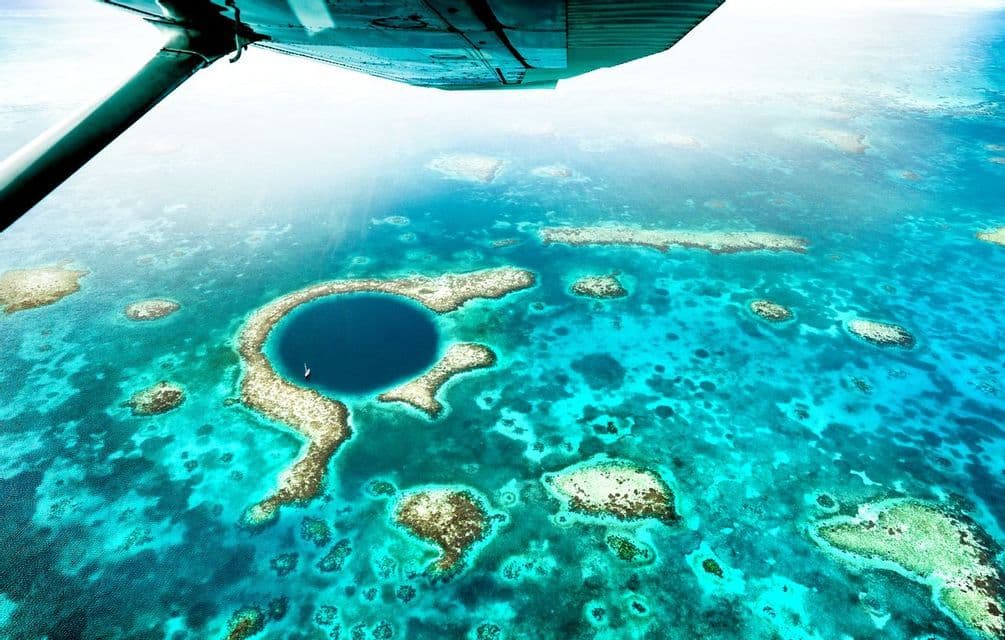An aerial view from under an airplane wing of a large blue hole surrounded by coral reefs in a turquoise ocean.