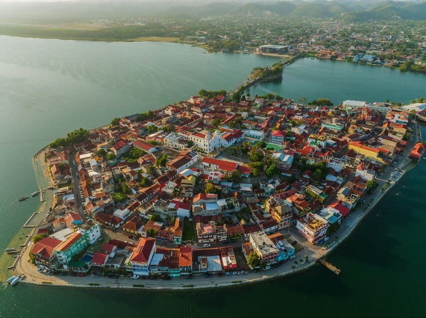 Aerial shot of a small city on a peninsula with colorful roofs, surrounded by calm water and connected by a causeway to the shore.