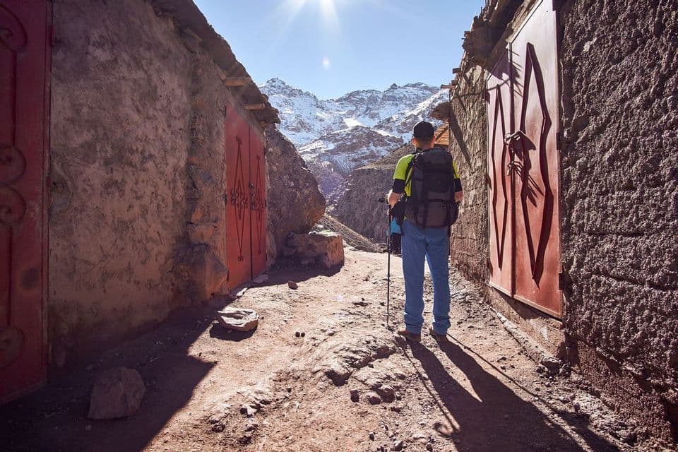 A hiker with a backpack and trekking poles stands on a dirt path between rustic buildings, facing a range of sunlit, snow-capped mountains.