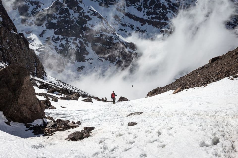 A WeRoad group trip hikes up a snowy trail in a rocky mountain valley under a partly cloudy sky.