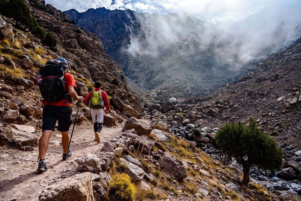 Two hikers from a WeRoad group trip, seen from behind, walk along a rocky mountain trail into a cloud-filled valley.