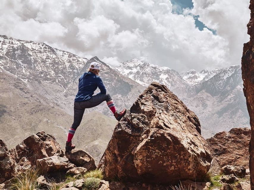 A hiker in a blue shirt and white cap stands on a rocky outcrop, looking out at a range of snow-capped mountains under a cloudy sky.