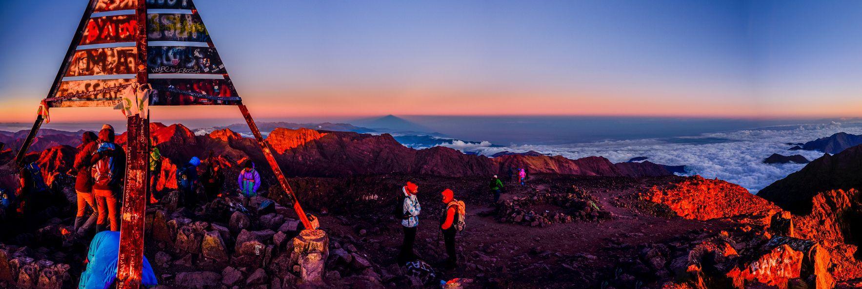 A WeRoad group trip on a mountain summit at sunrise, looking out over a sea of clouds and the peak's shadow on the horizon.