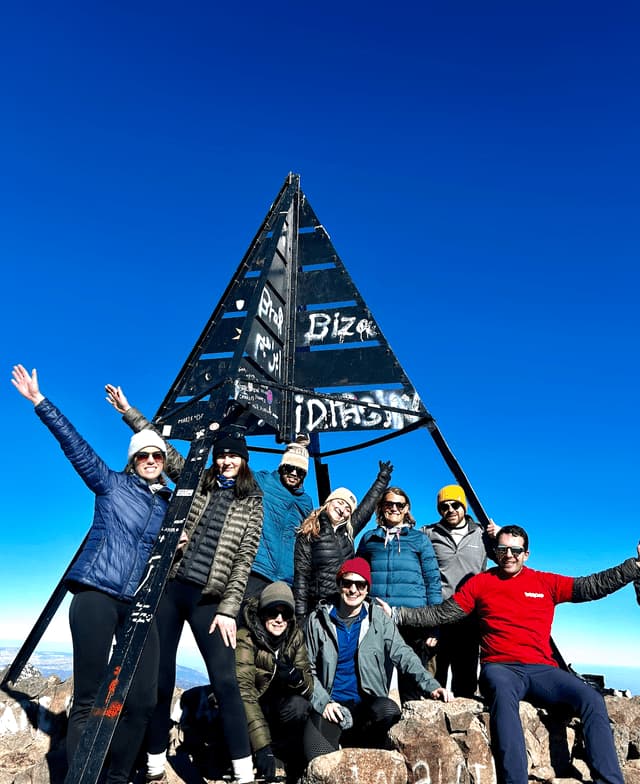 A WeRoad group trip posing on a rocky mountain summit next to a black metal pyramid marker under a clear blue sky.