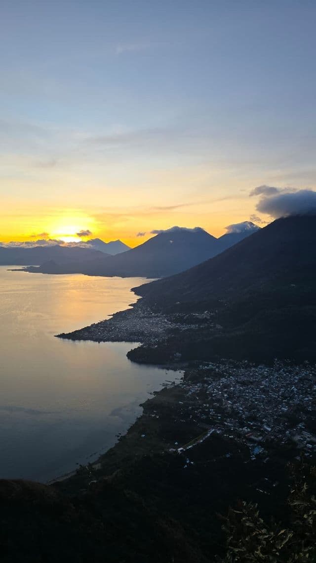 Vista elevada de un pueblo a orillas del lago rodeado de montañas al amanecer, con el resplandor del sol en el horizonte.
