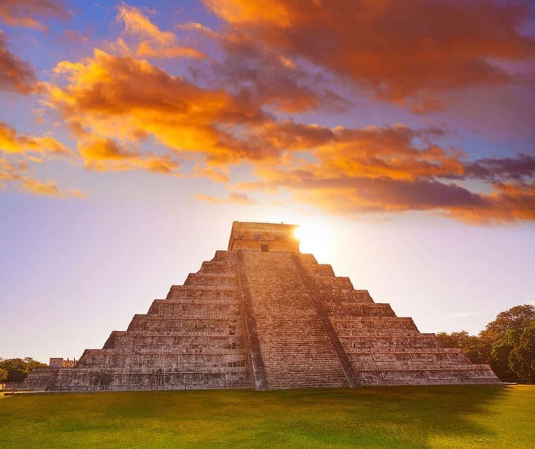 A large stone step pyramid on a green lawn with the sun setting behind it, casting a glow under a cloudy orange sky.