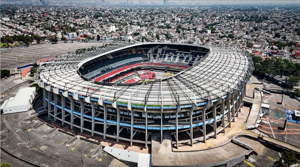 An aerial view of a large, empty football stadium with a complex roof structure, surrounded by a vast, sprawling cityscape.