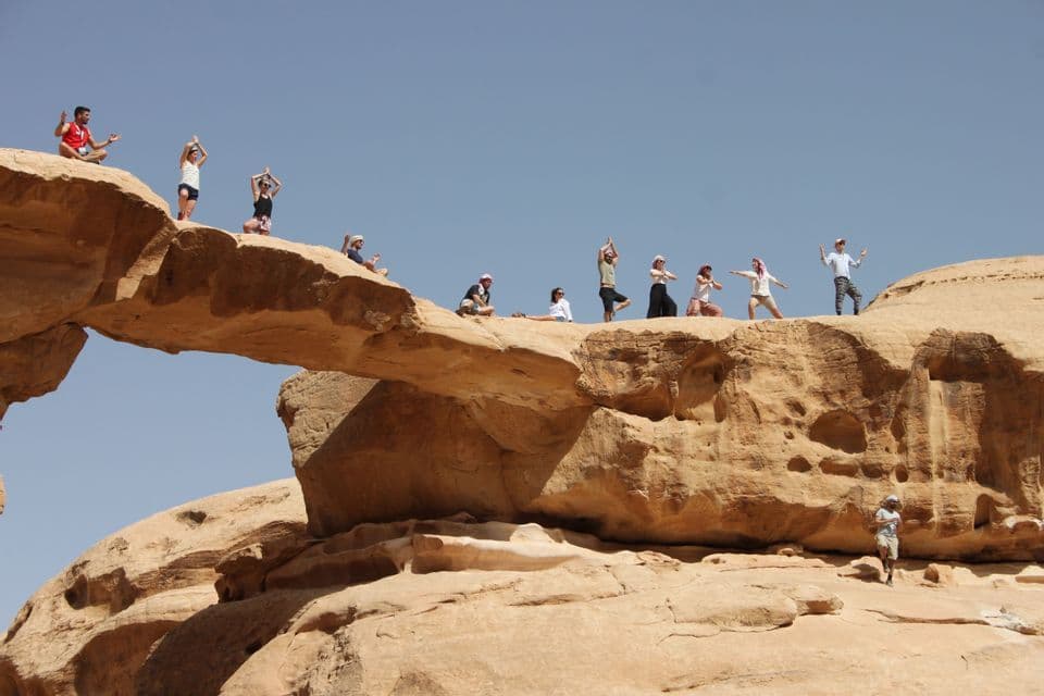 Eine WeRoad Gruppenreise, die auf einem großen Sandsteinbogen in der Wüste unter klarem blauem Himmel posiert und Yoga macht.