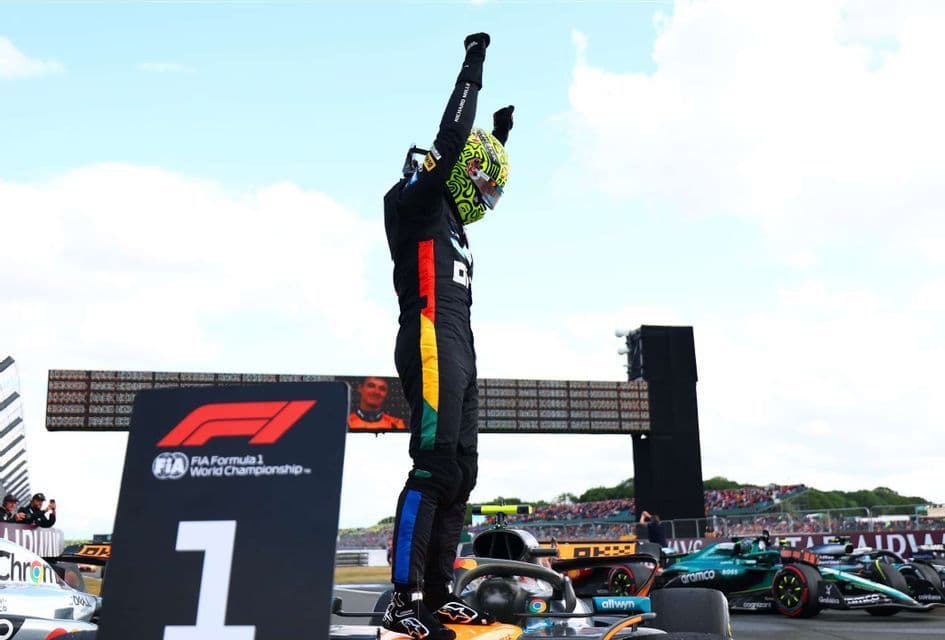 A Formula 1 driver in a black racing suit stands on his car with arms raised in victory next to a number one sign on the racetrack.