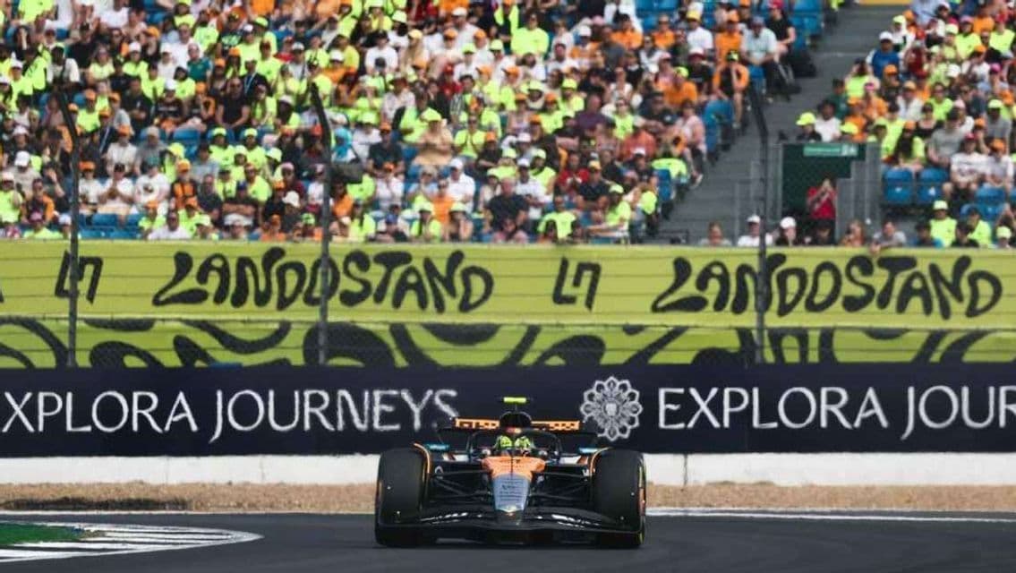 An orange and black Formula 1 car on a racetrack, with a crowd of spectators in a grandstand behind a 'LANDOSTAND' banner.