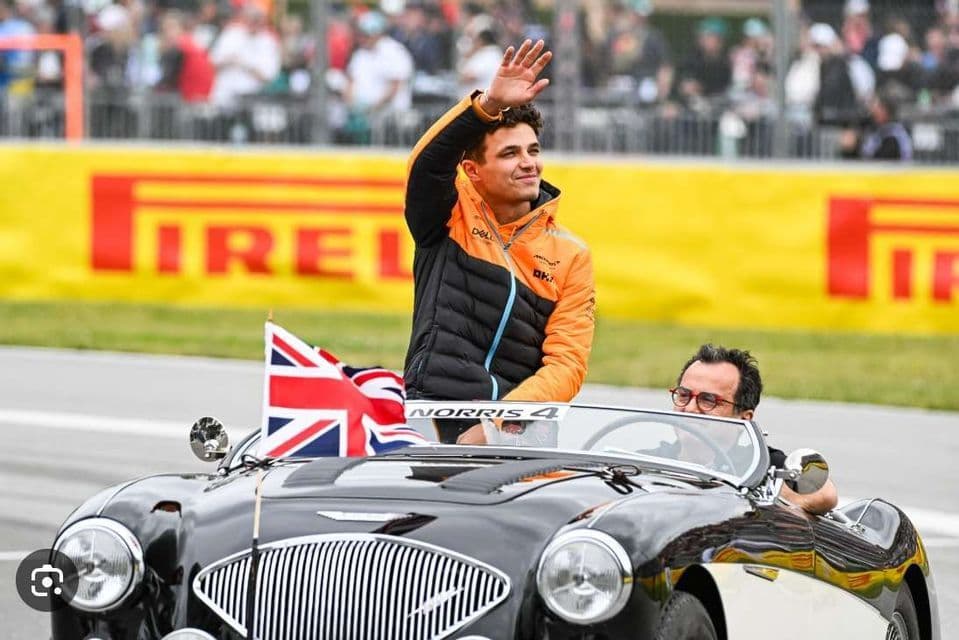 A man in an orange and black jacket waves to a crowd while standing in a classic black convertible car with a British flag.
