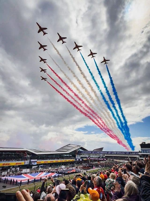 Nine jets fly in formation, leaving trails of red, white, and blue smoke over a crowded grandstand during an airshow.