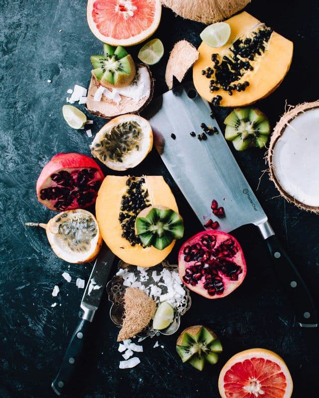 An overhead view of various sliced tropical fruits like papaya, kiwi, and pomegranate with two knives on a dark surface.