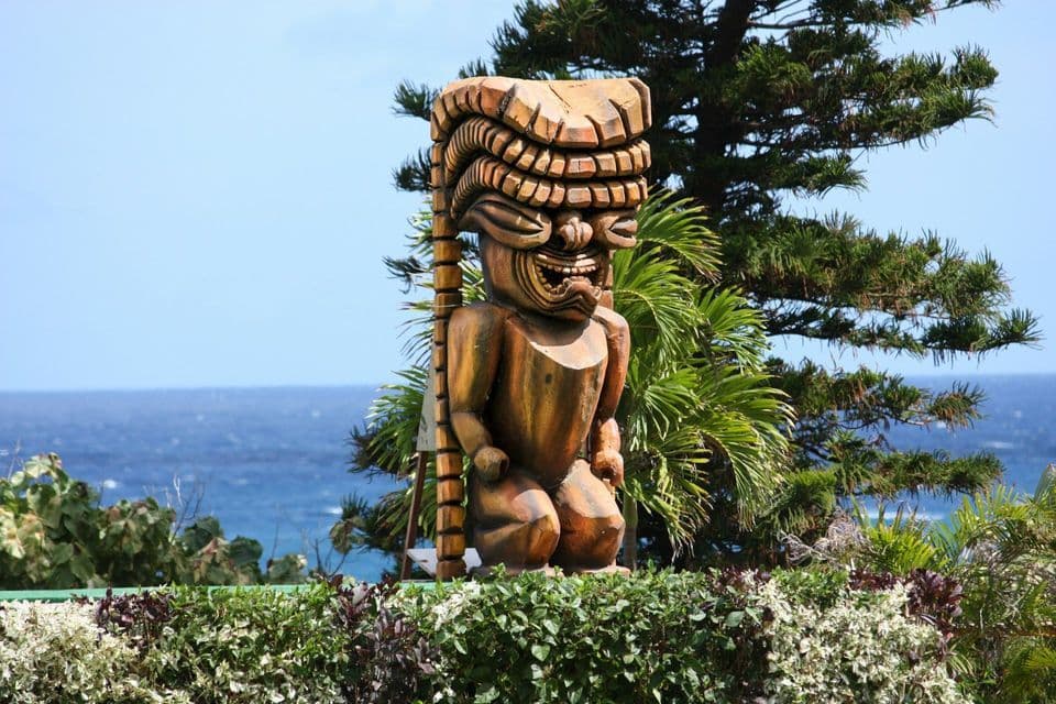 A carved wooden tiki statue with a detailed headdress stands among green plants, with a clear blue ocean in the background.