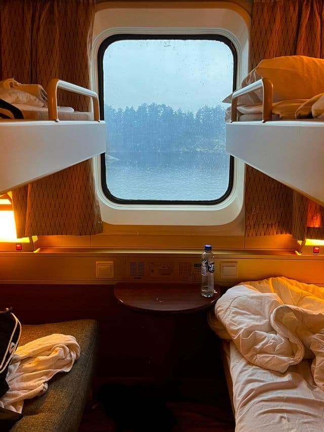 L'interno di una cabina di un treno letto con due letti a castello sfatti e una vista su un lago boscoso dalla finestra.