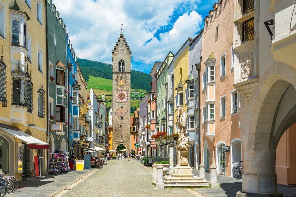A cobblestone street lined with colorful buildings leads toward a stone clock tower with a green hill visible in the background.