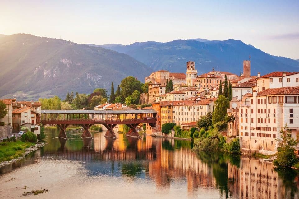 A covered wooden bridge spans a calm river in front of a historic town, with mountains in the background under a warm sky.