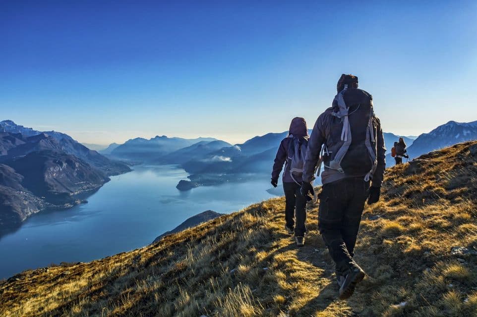 A WeRoad group trip hiking on a mountain trail, overlooking a vast lake and distant mountains under a clear sky.