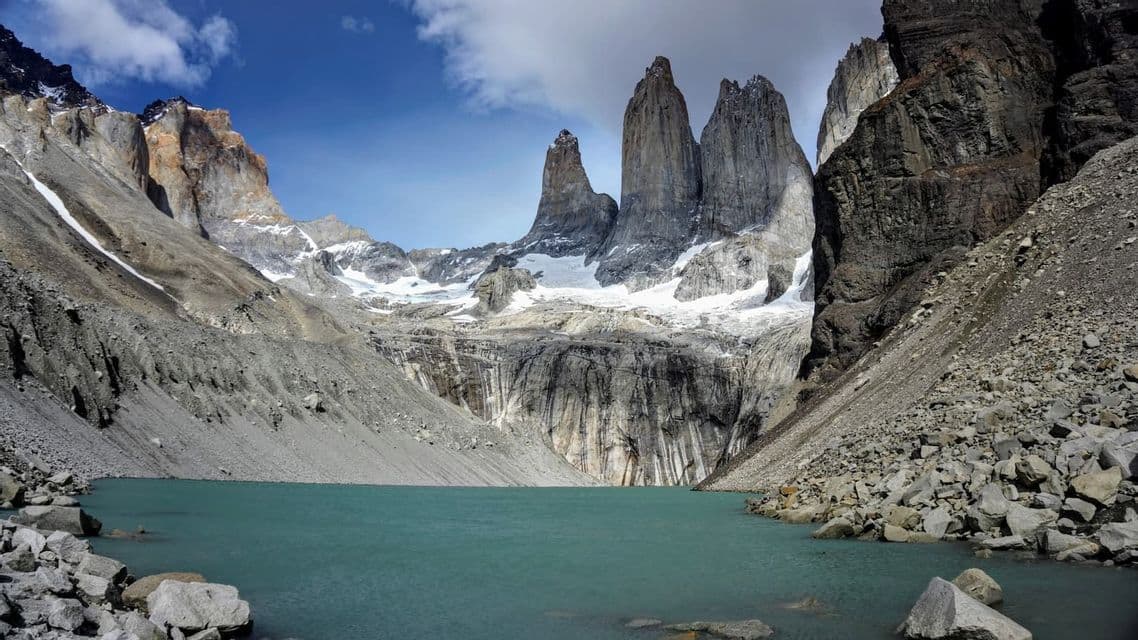 Un lago glaciale turchese ai piedi di imponenti guglie rocciose e ripidi ghiaioni, sotto un cielo azzurro con alcune nuvole.