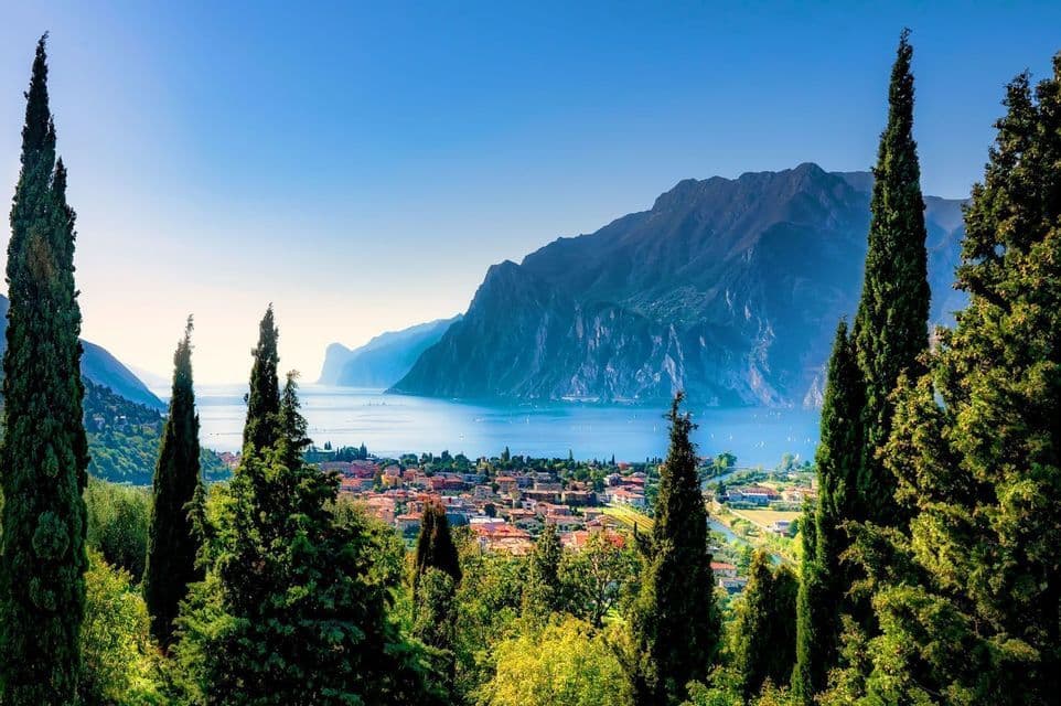A sunlit town on the shore of a large blue lake, with steep mountains rising in the distance, seen through tall green cypress trees.