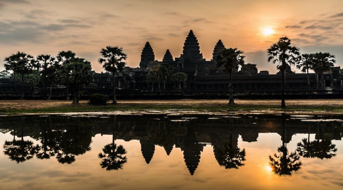 Ein alter Tempel mit mehreren Türmen zeichnet sich vor dem Sonnenaufgang ab und spiegelt sich in einem See mit Palmen.
