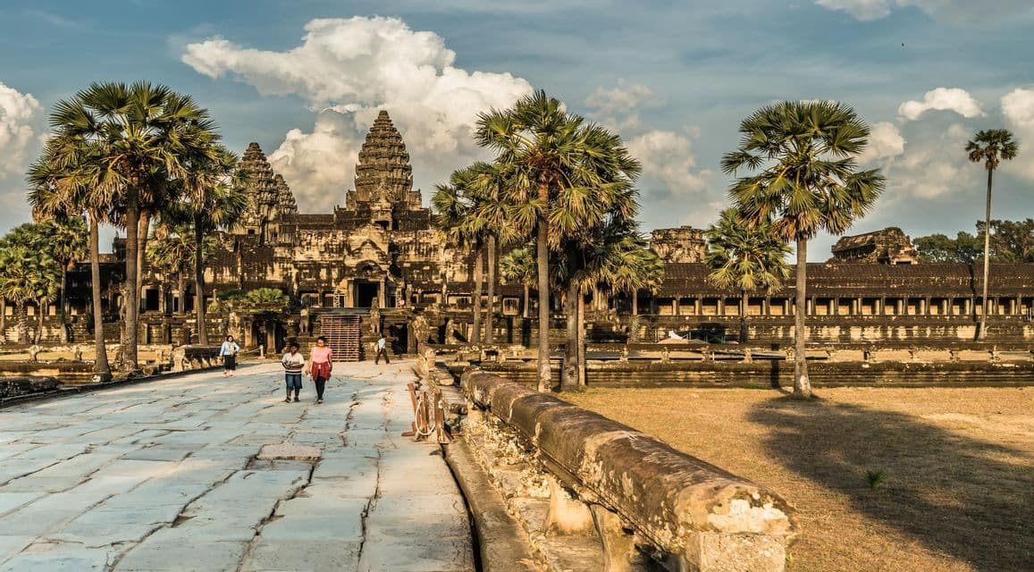 A few people walk along a wide stone causeway leading to a large, ancient temple complex flanked by tall palm trees under a partly cloudy sky.