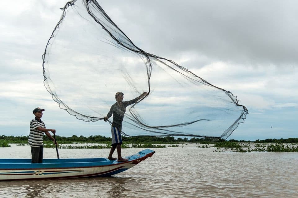 Dos pescadores en una pequeña barca en un río, uno de pie en la proa y lanzando una gran red de pesca.