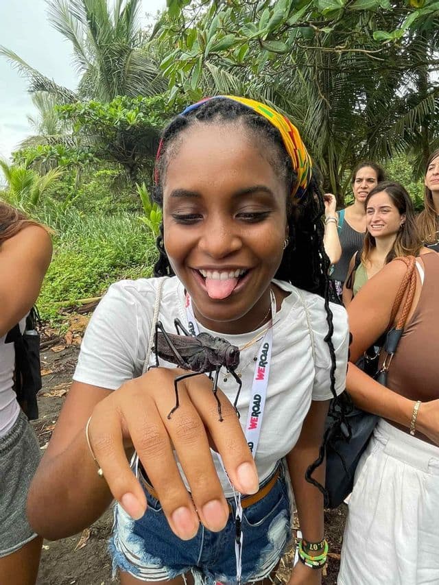 Una donna sorridente durante un viaggio di gruppo WeRoad tiene giocosamente un grande insetto nero sulla mano, all'aperto in un ambiente lussureggiante.