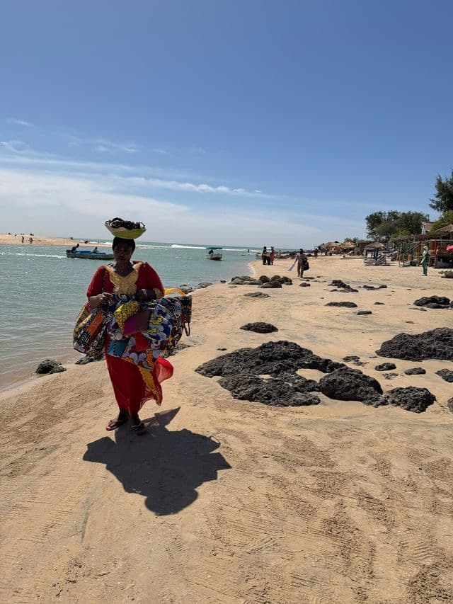 Una mujer con un vestido rojo camina por una playa de arena, equilibrando una cesta en la cabeza y llevando coloridas bolsas de tela.