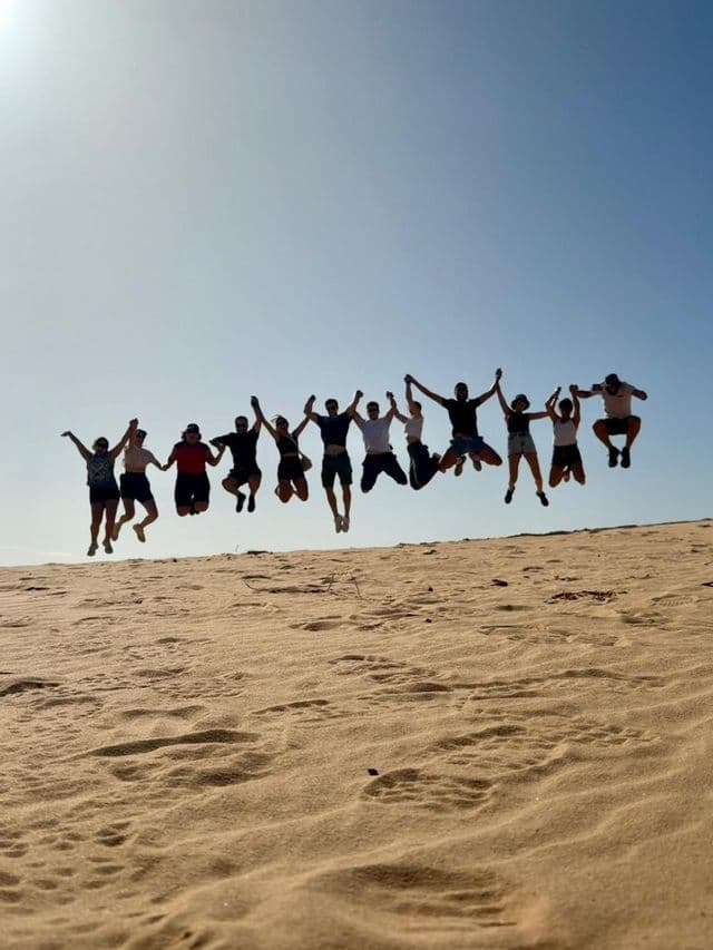Un viaje en grupo de WeRoad, cogidos de la mano y saltando juntos en el aire en una duna de arena bajo un cielo brillante y despejado.
