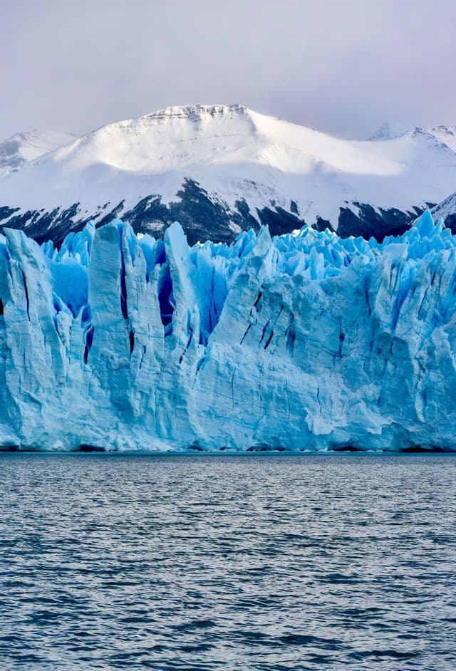 Un enorme ghiacciaio blu si erge dalle acque scure, con montagne innevate sullo sfondo.