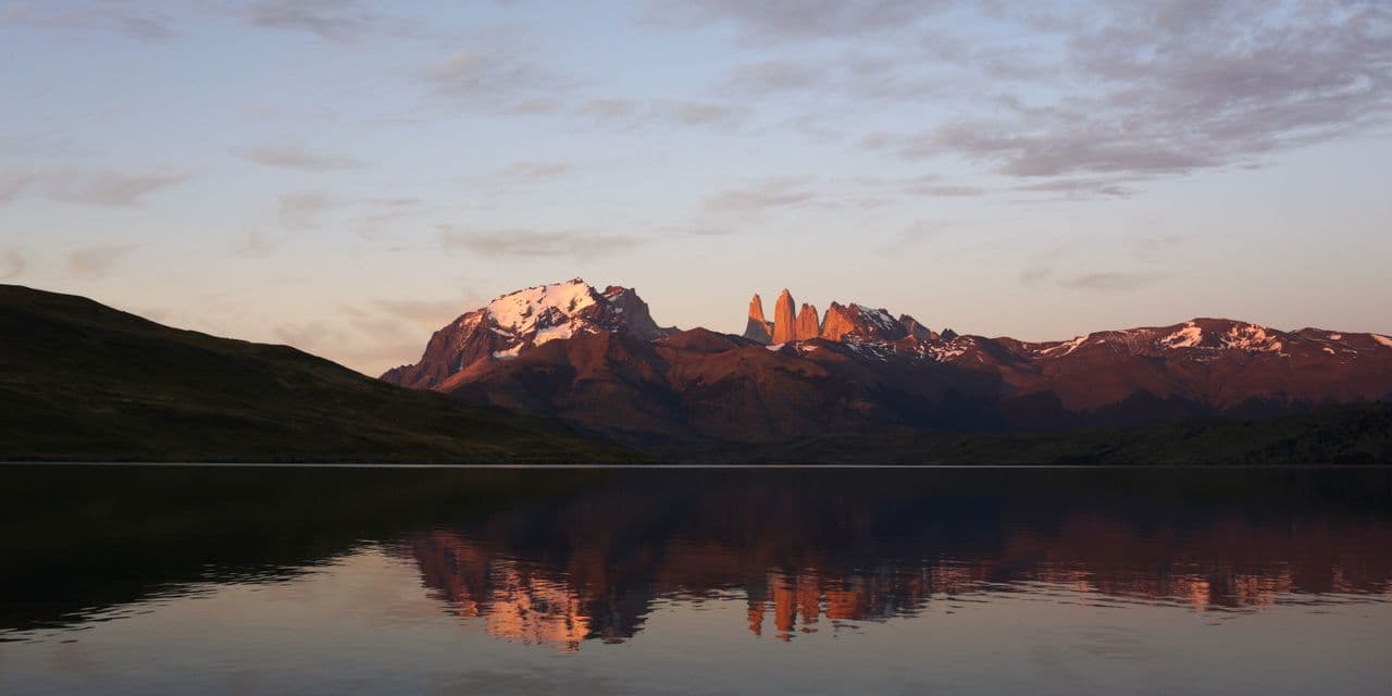 A snow-capped mountain range, with peaks glowing in the sunrise, is reflected in the calm waters of a large lake.