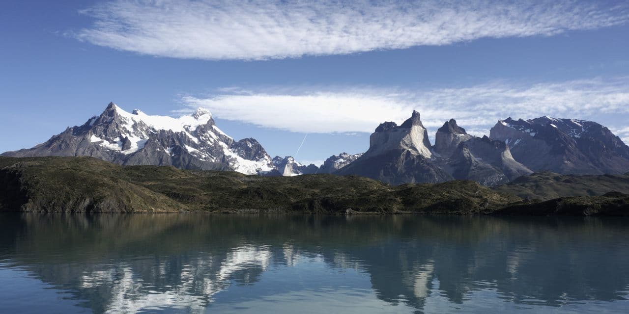 A wide shot of snow-capped and jagged mountains reflected in a calm lake, with green hills lining the shore.