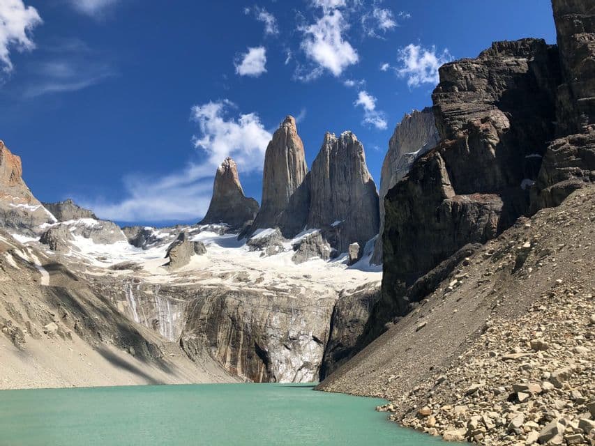 Three jagged granite peaks and a glacier towering over a turquoise glacial lake under a bright blue sky with sparse clouds.