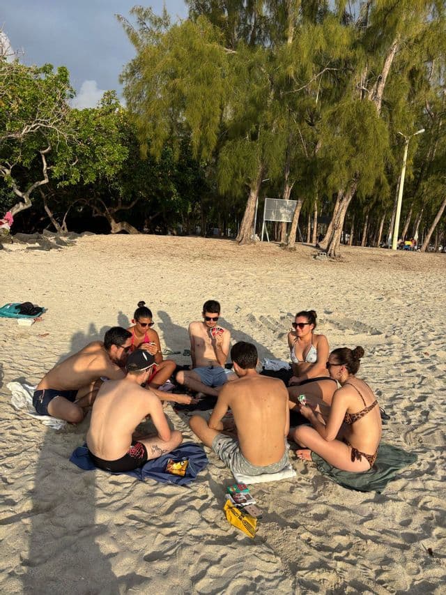 Un voyage de groupe WeRoad assis en cercle sur une plage de sable, jouant aux cartes ensemble à l'ombre des arbres.