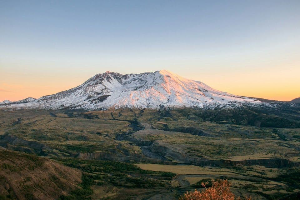 Una cima montuosa innevata è illuminata dall'alba, con una vasta valle che si estende sotto un cielo sereno.