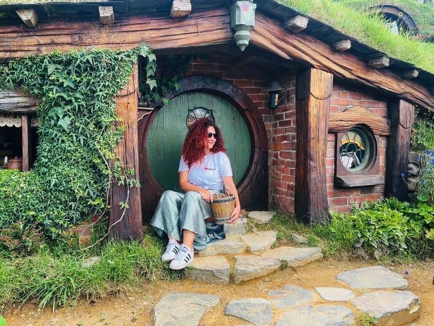 Une femme aux cheveux roux bouclés est assise sur les marches en pierre devant une maison avec une porte ronde verte et un toit végétalisé.