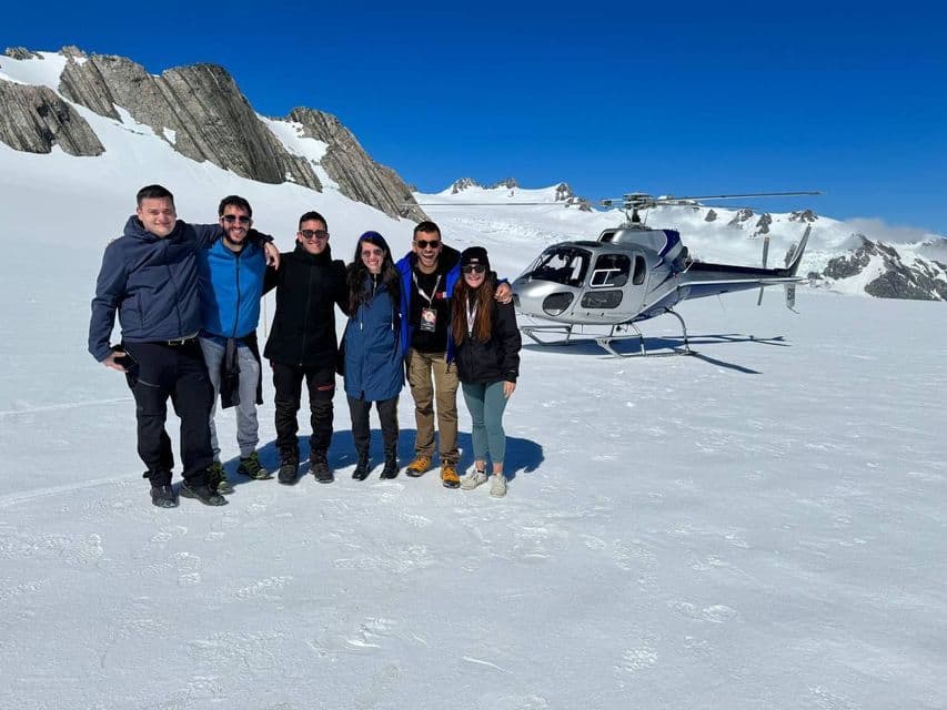 Eine WeRoad Gruppenreise von lächelnden Menschen auf einem verschneiten Gletscher mit einem Helikopter und Bergen im Hintergrund.
