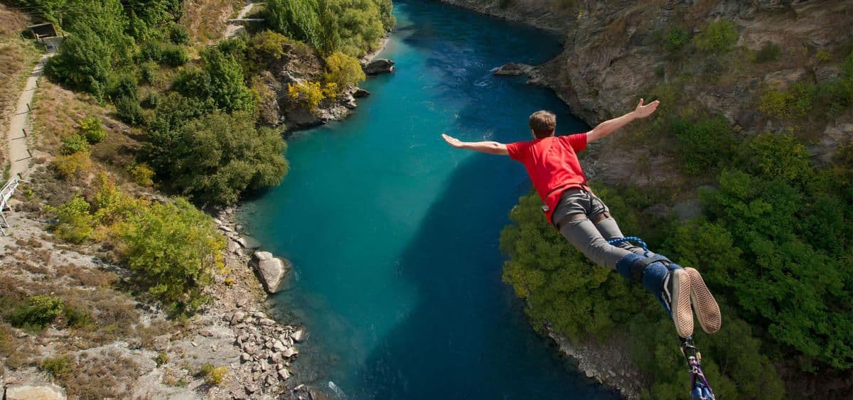 Une personne en t-shirt rouge faisant du saut à l'élastique les bras tendus au-dessus d'une rivière turquoise dans un canyon rocheux.
