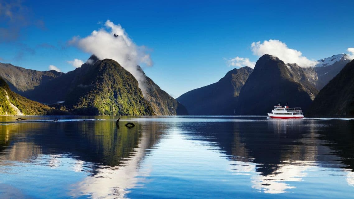 Un bateau touristique rouge et blanc navigue sur des eaux calmes à travers un fjord, des montagnes vertes et abruptes se reflétant à la surface.